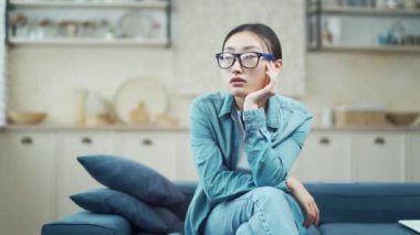 Sad young asian woman sitting with head on hand on sofa in living room. Upset female in glasses and casual clothes thinking about her problems indoors She is tired, unhappy, disappointed and depressed