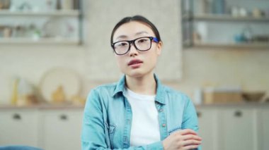 Close-up portrait of young confident asian woman with crossed arms looking at camera indoors. Head shot beautiful serious female with calm face wearing glasses and casual clothes posing in living room