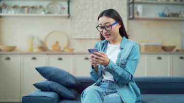 Young smiling asian woman using smartphone while sitting on sofa in living room. Happy female holding mobile phone texting, shopping online or browsing social media on cosy couch in kitchen at home