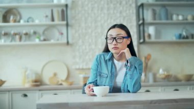 Sad young asian woman sitting at the kitchen table at home. Upset female in glasses holds a cup of coffee and thinks about her problems indoors. She is tired, unhappy, disappointed and depressed