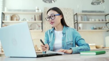 A smiling young asian woman is chatting online using a laptop and writing in a notebook while sitting at the kitchen table Female freelancer or student in glasses waving and cheerfully talking at home