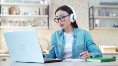 Young asian woman wearing headphones typing on laptop and writing in notebook while sitting at desk in living room. Freelancer or student in glasses browsing the internet at home in the kitchen