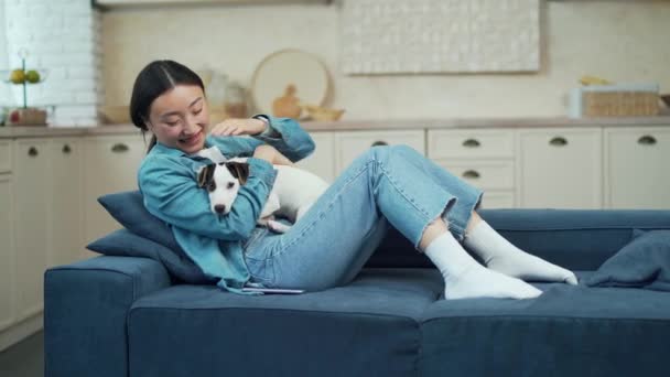 リビングルームのソファに横に小さな白と黒の犬を保持する幸せな若いアジアの女性 笑顔で明るい女性は喜んでストロークし 自宅でソファの上に面白いペットと遊んでいます — ストック動画