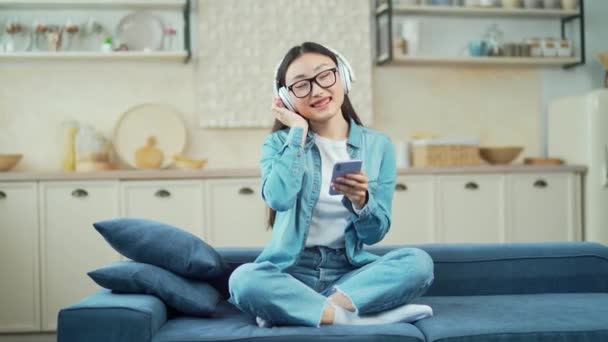 一位身穿耳机 手持智能手机的年轻亚洲女人坐在客厅的沙发上听音乐 一个笑着闭上双眼的女人在家里的沙发上放松一下 — 图库视频影像
