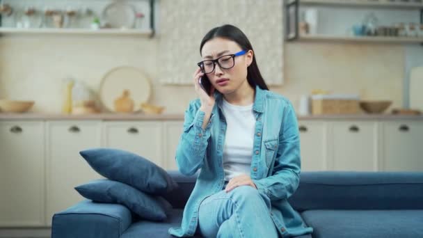 リビングルームのソファに座っている間 若いアジアの女性は電話で頭痛の種の話をしています メガネとカジュアルな服を着た疲れた女性は スマートフォンで話している間に頭のマッサージを与えます — ストック動画