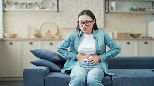 年轻的亚洲女人坐在客厅的沙发上 感到胃部紧张疼痛 一个穿着休闲装的女人在家里有一个不舒服的胃 急性胃炎 抽筋一种抽筋或心痛的感觉 — 图库视频影像