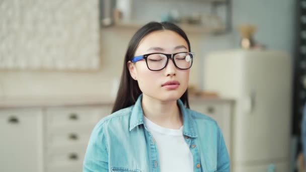 リビングルームに座っている間に目を閉じて瞑想若いアジアの女性のクローズアップ 自宅で深呼吸した後にリラックスしたカジュアルな服に眼鏡をかけた女性の肖像画 — ストック動画
