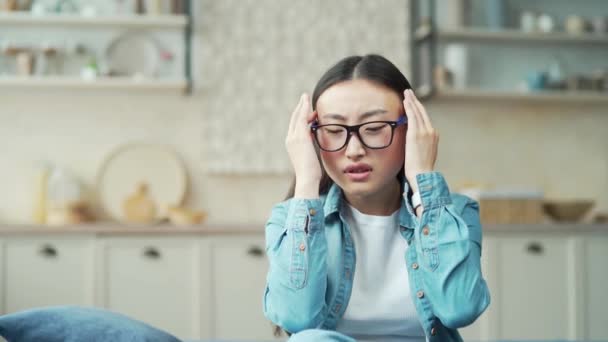 年轻的亚洲女人坐在客厅的沙发上感到头疼的特写镜头 疲惫的女人戴着眼镜 穿着休闲装 闭着眼睛 头和鼻梁都在按摩 — 图库视频影像