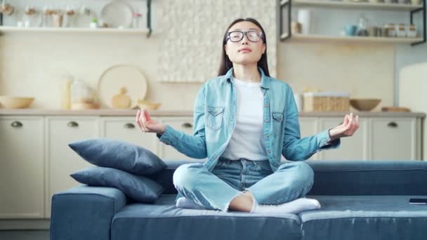リビングでソファーに座りながら目を閉じて瞑想する若いアジア人女性 家庭で快適なソファの上に深く呼吸蓮の位置でリラックスカジュアル服の女性 — ストック動画