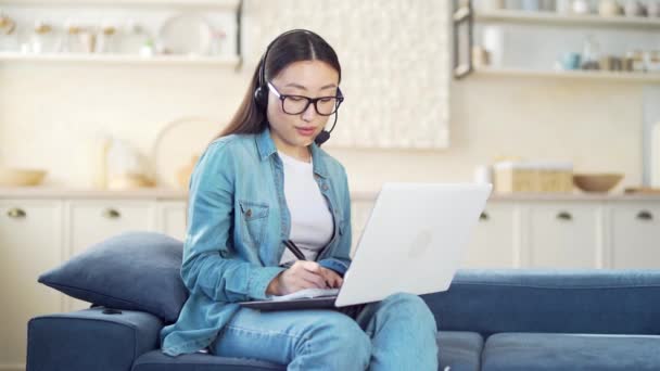 ヘッドセットとラップトップを持つ若いアジアの女性は オンラインで通信し リビングルームのソファに座っている間にノートブックに書き込みます 女性学生ビデオ呼び出しを介して家庭でソファ屋内 — ストック動画