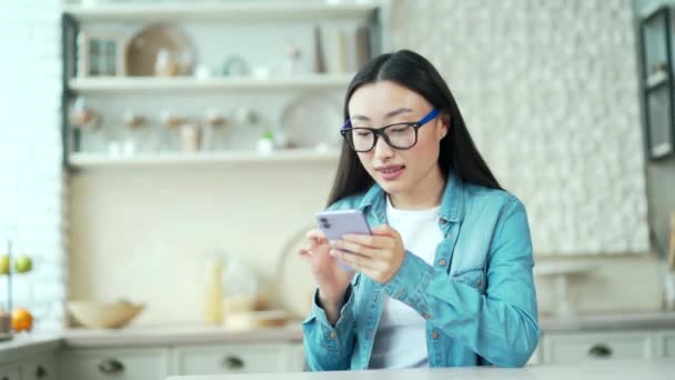 快乐的年轻亚洲女人坐在厨房里 一边用智能手机在线聊天 戴眼镜 面带微笑的女性在网上购物或浏览社交网站 手里拿着小玩意 — 图库视频影像