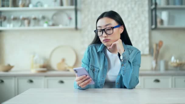 Sad Young Asian Woman Browsing Smartphone While Sitting Kitchen Table — Vídeos de Stock