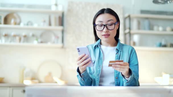 快乐的年轻亚洲女人坐在家里的桌子边 一边用智能手机输入信用卡号码 戴眼镜 面带微笑的女人上网购物 手里拿着小玩意和信用卡 — 图库视频影像