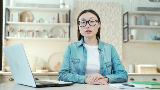 自宅のノートパソコンの近くの机に座ってカメラを見ている若いアジア人女性の肖像職場の屋内でカジュアルな服の中でガラスを身に着けている深刻なかなり穏やかな女性のフリーランスや学生 — ストック動画