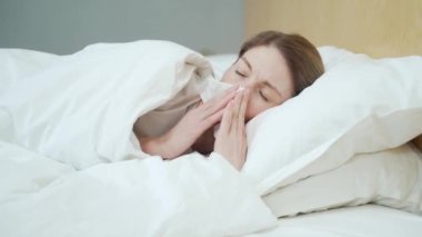 Sick blond woman blowing running nose sneezing and coughing laying on the warm bed in the bedroom Unhealthy female under the blanket getting flu virus symptom Cold and fever concept