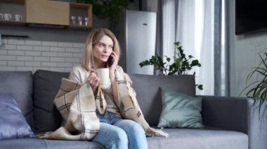 Sıcak çay bardağıyla kanepede, sıcak battaniyenin altında oturan hasta sarışın kadın. Evde şiddetli baş ağrısı var. Sağlıksız bir kadın grip virüsü belirtileri gösteriyor.