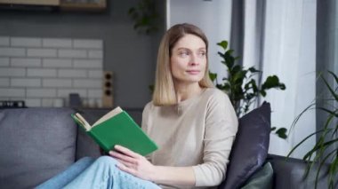 Close-up. Mature beautiful woman reading a book while sitting on the sofa in the living room at home. Relaxing on a comfortable couch during free time on weekends. Happy female student with a textbook