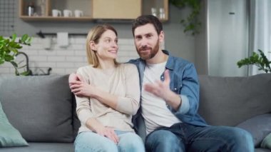 Happy man and woman looking at camera, waving and chatting online while sitting on sofa in living room at home. A cheerful family couple is hugging and talking via video link with relatives or friends