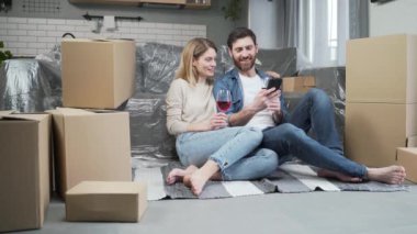A happy married couple is celebrating a housewarming. Adult man and woman barefoot sitting on the kitchen floor in a new apartment and drinking wine in the middle of cardboard boxes They hug and smile