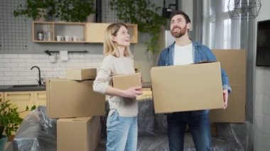 A happy married couple moves into a new apartment. Adult joyful man and woman enter the kitchen, holding cardboard boxes in their hands. They are satisfied, look around the room, hug and smile