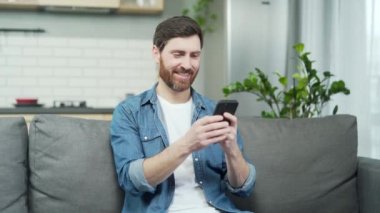 Bekar sakallı bir adam, flört uygulamasında kadın profillerini karıştırıyor ve oturma odasında akıllı telefondaki fotoğrafları beğeniyor ya da beğenmiyor. Sosyal platformda partner arayan ve eşleşme bekleyen genç bir adam.