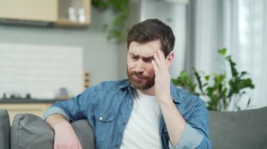 Mature handsome man feels headache while sitting on couch in living room. A bearded middle-aged male in casual clothes is massaging his temples with his hand at home. He is tired and has a migraine
