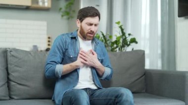Man having heart attack sitting alone at home on the sofa. Mature bearded Caucasian male holding chest and breathing heavily in room. Indoor Suffering from chest pain on sofa