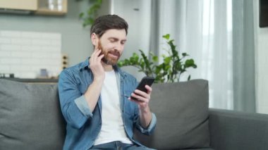 Mature handsome man feels toothache while sitting on couch in living room at home. A bearded middle aged aucasian male in casual clothes is bothered by a sore tooth while texting on a smartphone