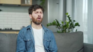 Sick bearded man suffers from seasonal allergies blowing running nose sneezing and coughing sitting on couch in living room at home Unhealthy male shirt getting flu virus symptom Cold and fever indoor