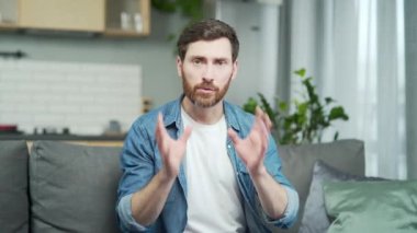 Mature caucasian man looking at camera and talking while sitting on sofa at home. Webcam view of confident bearded male having conversation with virtual meeting interlocutor, gesturing with hands