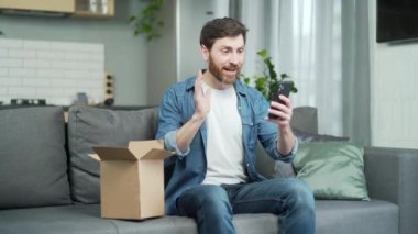 Happy handsome man chatting online on smartphone thanks friend for received gift sitting on couch at home. Joyful malethanks for the parcel, which he is very pleased with. Satisfied client concept