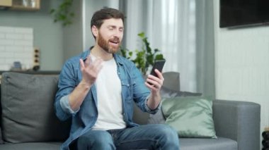 Evdeki oturma odasında kanepede otururken elinde akıllı telefonla sohbet eden mutlu ve olgun bir adam. Neşeli sakallı beyaz erkek arkadaşına el sallıyor, neşeli bir şekilde el sallıyor, gülüyor,