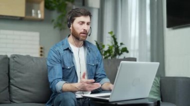 A man in headphones using a laptop computer communicates via video chat. Male waving hands having e-meeting work remotely in home. Webinar, videocall event, video conference call communication