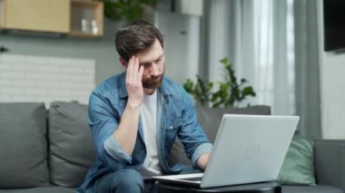 Serious mature handsome man feeling headache at workplace at home working on laptop while sitting on sofa. Overtired freelancer is massaging his temples with his hands. Middle-aged male has a migraine