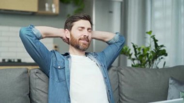 Mature handsome man stretching while sitting on sofa at home. A middle-aged man puts his hands behind his head, closes his eyes and smiles. A bearded freelancer relaxes and unwinds after the work done
