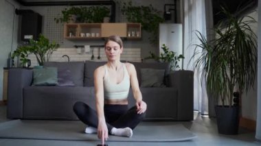 Calm relaxed woman sitting in lotus position meditating and breathes slowly while make yoga asanas in the living room Athletic female meditation practice using smartphone at home