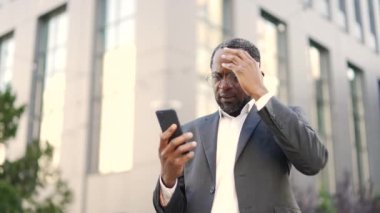 Afro-Amerikalı işadamı dışarıda akıllı telefonu tutarken şok edici bir mesaja bakıyor. Takım elbiseli olgun yatırımcı ofis binasının önünde kötü haberler okuyor.