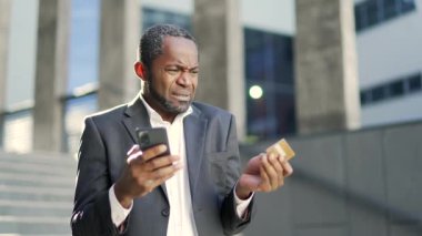 Üzgün Afro-Amerikalı yetişkin bir adam dışarıda dururken elinde kredi kartı ve akıllı telefon tutuyor. Dolandırıcılıkla yüzleştiği için kızgın. Depresyondaki erkek hesabından para çalındığını fark etti.