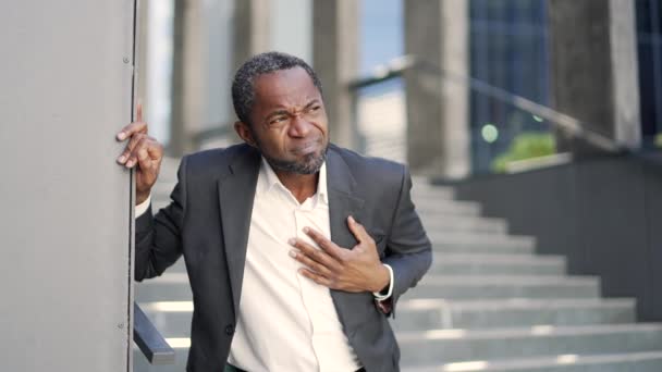 病重的非洲裔美国成年人 心脏病缠身 站在办公楼前 一个穿着正式西服的中年商人把他的手放在胸前 他心脏病发作了 — 图库视频影像