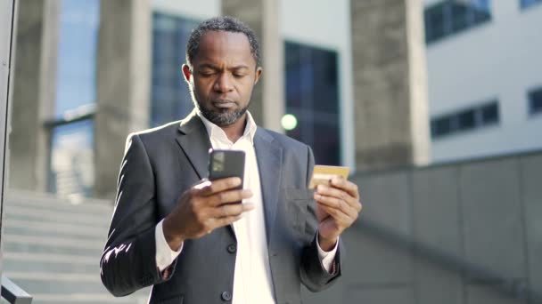 非洲裔美国人成熟男子在智能手机上输入信用卡号码 同时站在办公楼前 穿着西装的商人笑了 因为他在网上买得很成功 — 图库视频影像