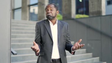 Şaşırmış afro-amerikalı yetişkin adam ellerini açar, omuz silker ve dışarıda dururken kameraya bakar. Resmi takım elbiseli bir erkeğin yüzünde karışık bir ifade vardır. Şüphe kavramı