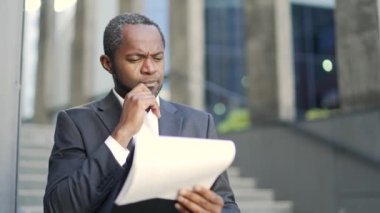 Ciddi bir Afro-Amerikalı yetişkin adam ofis binasının önündeki bir belgeyi inceliyor. Resmi takım elbiseli konsantre bir işadamı elinde bir iş mektubu tutuyor ve bir sorun hakkında düşünüyor.