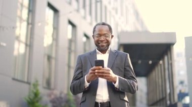 Gözlüklü, gülümseyen Afro-Amerikan iş adamı dışarıda dururken e-postaları kontrol ediyor. Takım elbiseli mutlu bir yatırımcı ofis binasının önünde iyi haberler okumaktan mutluluk duyar.