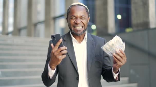 快乐的微笑着的非洲裔美国人 成熟的美国人 在一幢办公楼前拿着一大把现金钞票 兴奋地看着镜头 享受着自己的收入 — 图库视频影像