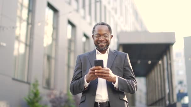 站在外面拿着智能手机 戴着眼镜检查邮件的非洲裔美国商人 穿着西装的快乐成熟的投资人在写字楼前看到好消息会很开心 — 图库视频影像