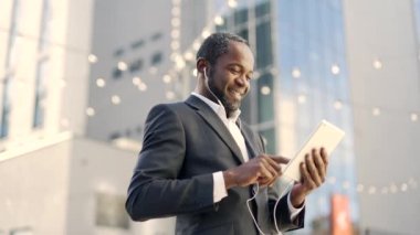 Dışarıda dururken kulaklıkla gülümseyen Afro-Amerikan iş adamı. Resmi takım elbiseli olgun iş adamı ofis binasının önünde internetten mesaj atarken iyi haberler okuyor.
