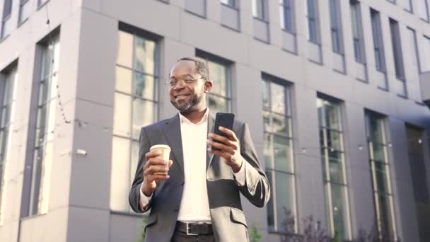 ガラスの中でアフリカ系アメリカ人のビジネスマンを笑顔にし コーヒーを飲みながら外を歩いているとスマートフォンを見ています オフィスビルの前で陽気にニュースを見てスーツの幸せな成熟した男 — ストック動画