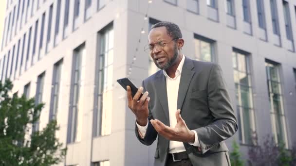 兴奋的非洲裔美国商人一边站在外面一边拿着智能手机一边感情用事 一个穿着正式西装的愤怒的成熟男人在一幢办公楼前的电话里吵架 — 图库视频影像