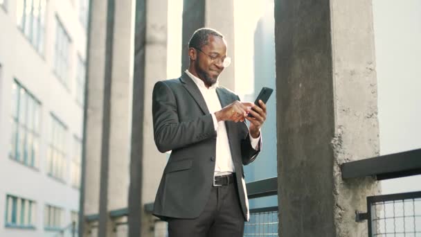 一位戴着眼镜 面带微笑的非洲裔美国商人站在街上用智能手机打字 穿着正式西装的快乐而成熟的男人在一幢办公楼前愉快地网上聊天 — 图库视频影像