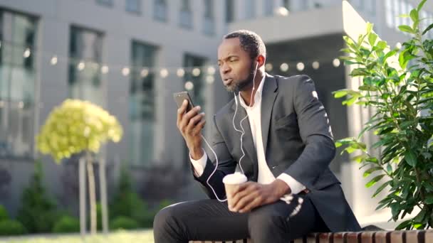 非洲裔美国人 头戴耳机 坐在长椅上 一边用智能手机说话 穿着西装 拿着一杯咖啡的商人正在一幢办公楼前谈话 — 图库视频影像